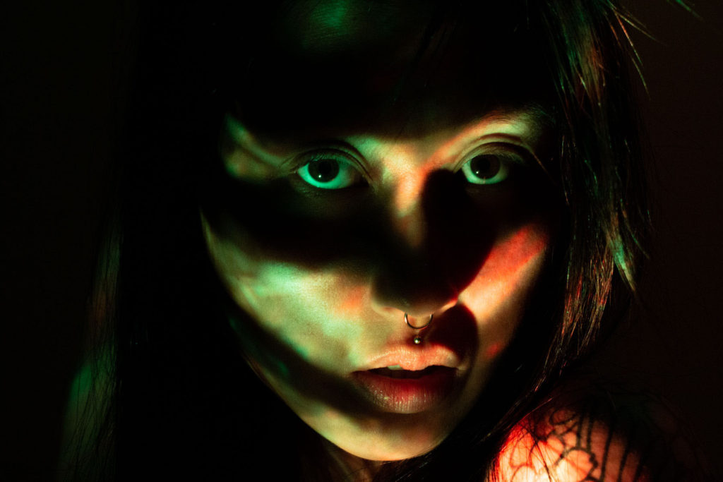 A self portrait photograph of Elaine. Her expression is neutral. Her face is slightly lit up with green and red lighting, however, there are some mysterious shadows casted over her face. The shadows look a bit like a mask over her eyes. She’s looking directly into the lens and both eyes are visible. 